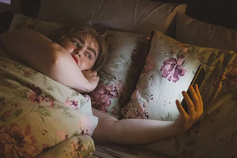 Maggie May: a woman in bed lit by her phone screen among floral pillows