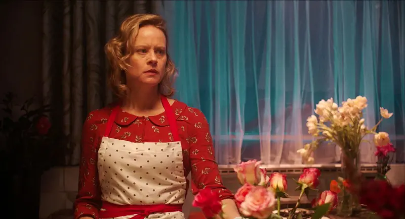 Mrs McCutcheon: a woman in a red dress and polka-dot apron stands by a window with roses