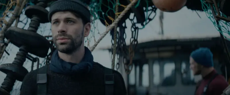 Never Never Never: a bearded fisherman in a beanie stands among nets and floats on the harbour