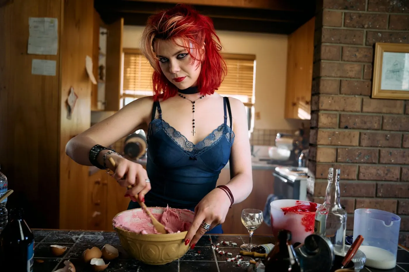 Penny Lane Is Dead: a red-haired young woman in a blue camisole mixes pink icing in a cluttered 1980s kitchen