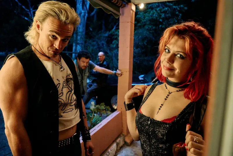 Penny Lane Is Dead: a bleached-blond man in a leather vest and a red-haired young woman on a porch at night, with men behind them